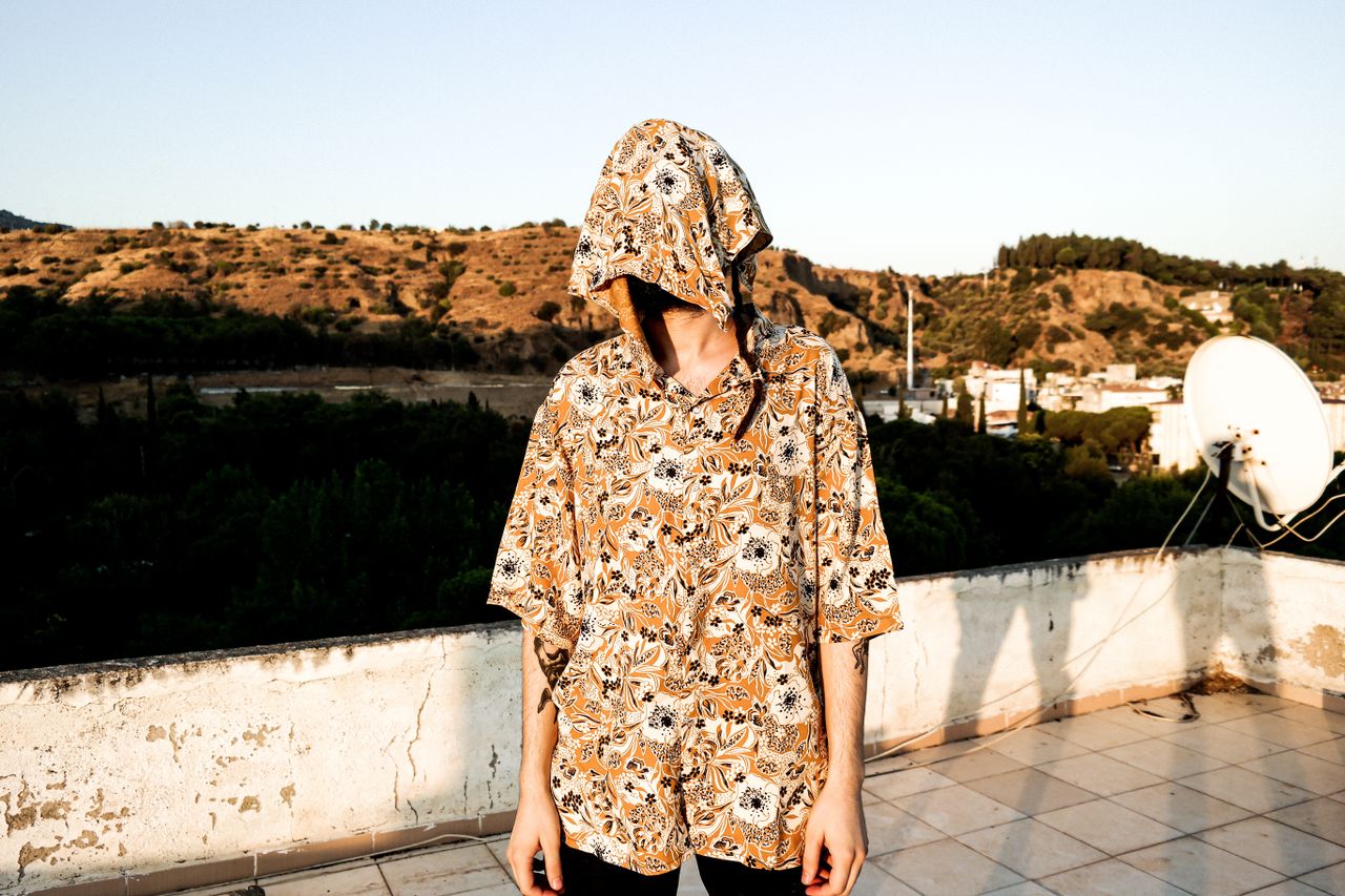 About Person in floral shirt on a rooftop, face covered, with a scenic hill backdrop.